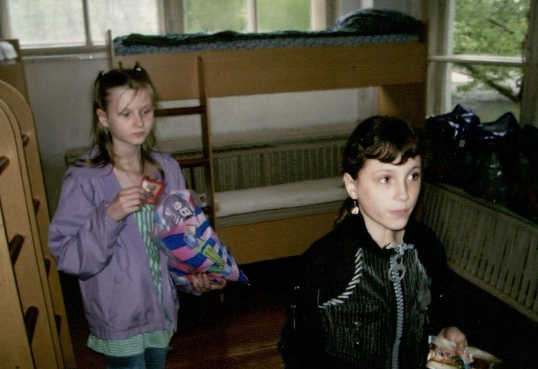 Before leaving for America, Masha paid the orphanage one last visit. While there, Masha shared desserts and treats with her friends, who scrounged around for last-minute gifts to give her. Masha is seen here in the bedroom she shared with the other orphan girls. Her friend Vica is in this picture. Masha slept in the top bunk pictured behind her in the photo by the window. She is holding some American candy that she will gift to her friends.